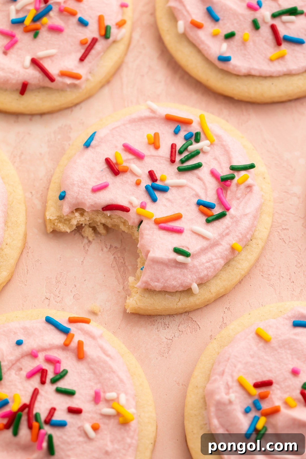Gluten-Free Cut-Out or Drop Sugar Cookies 3 Rows of gluten free sugar cookies topped with pink buttercream frosting and rainbow jimmies on parchment paper. One bite is missing from the cookie in the center of the photo.