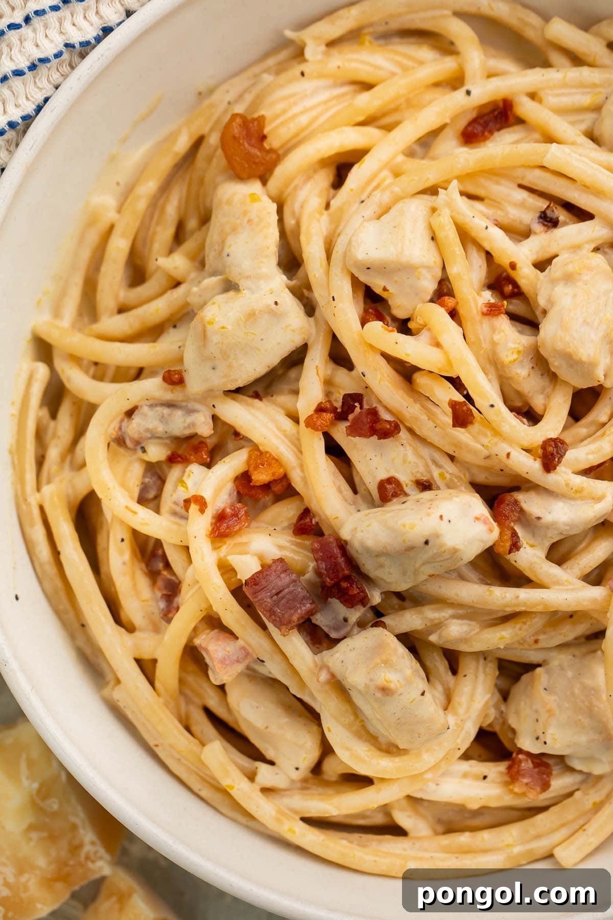 Close-up of a large white bowl of creamy chicken carbonara on a table with a blue and white kitchen towel, showing the rich texture and garnishes.