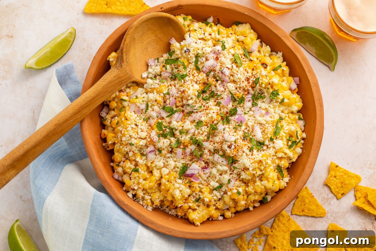 Top-down photo of a large wooden bowl holding a Mexican street corn salad, with fresh lime wedges and cilantro sprigs.