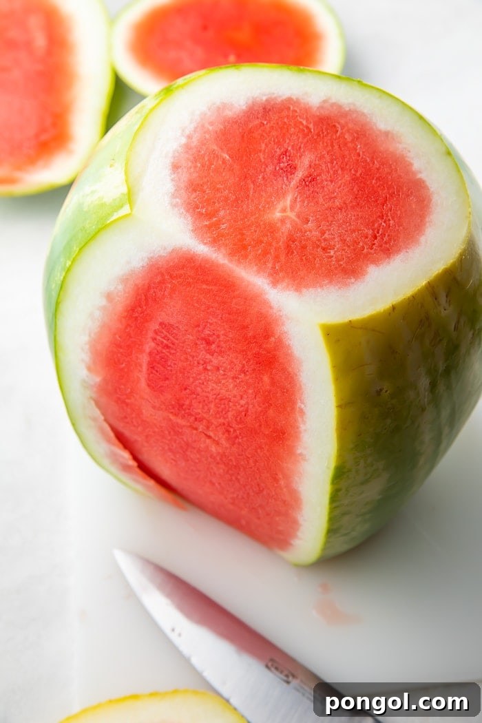 A watermelon standing upright with several strips of green rind already removed, exposing the bright pink flesh.