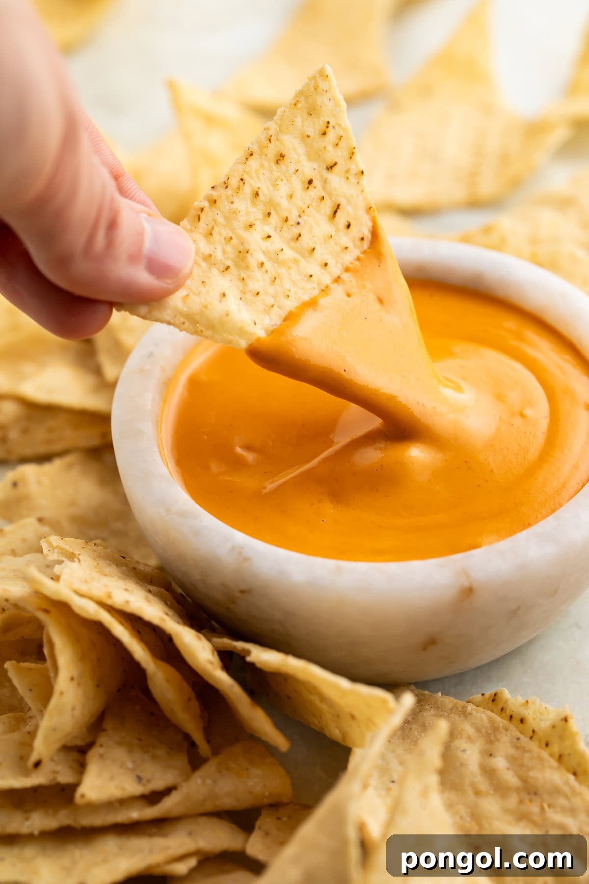 A woman's hand gracefully dips a crispy white corn tortilla chip into a pristine white bowl overflowing with vibrant orange vegan nacho cheese sauce.