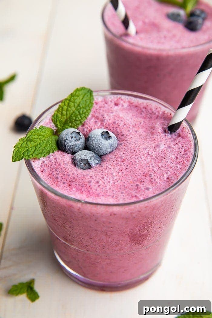 A vibrant blueberry smoothie in a tall glass, garnished with fresh blueberries and a straw, set against a bright, clean background.