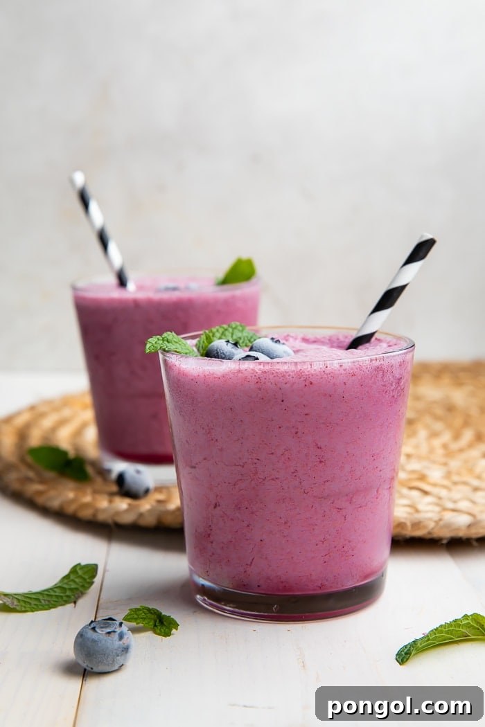 Two glasses of fresh blueberry smoothies, garnished with mint leaves and fresh blueberries, ready to be served on a light wooden surface.