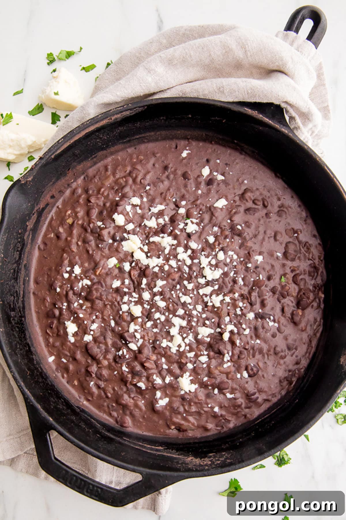 Refried black beans garnished with cotija crumbles in a large black cast iron skillet with a neutral cloth napkin around the handle, ready to be served.