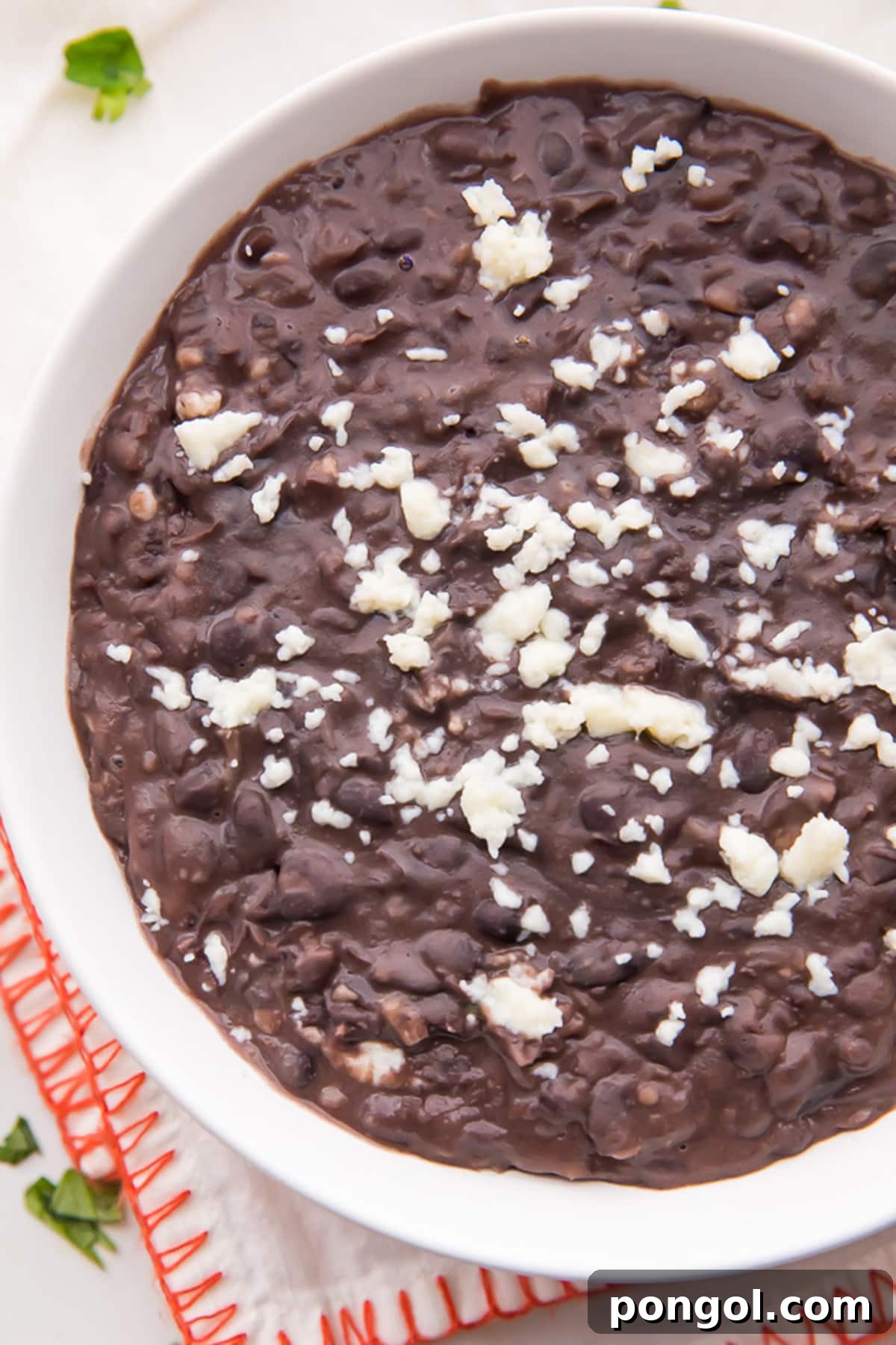 Refried black beans topped with queso fresco crumbles in a large white bowl resting on a red-edged cloth napkin.