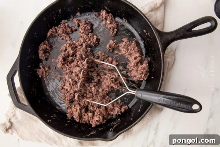 Mashed black beans in a large black cast-iron skillet, showing the continued mashing process.