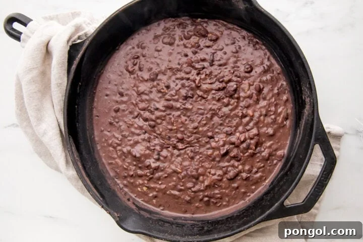 Refried black beans simmering in a cast-iron skillet, showing the addition of liquid.