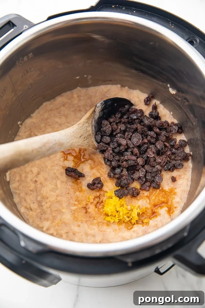 Overhead shot of freshly cooked rice pudding in an Instant Pot, with additional maple syrup, plump raisins, and aromatic lemon zest being stirred in with a sturdy wooden spoon