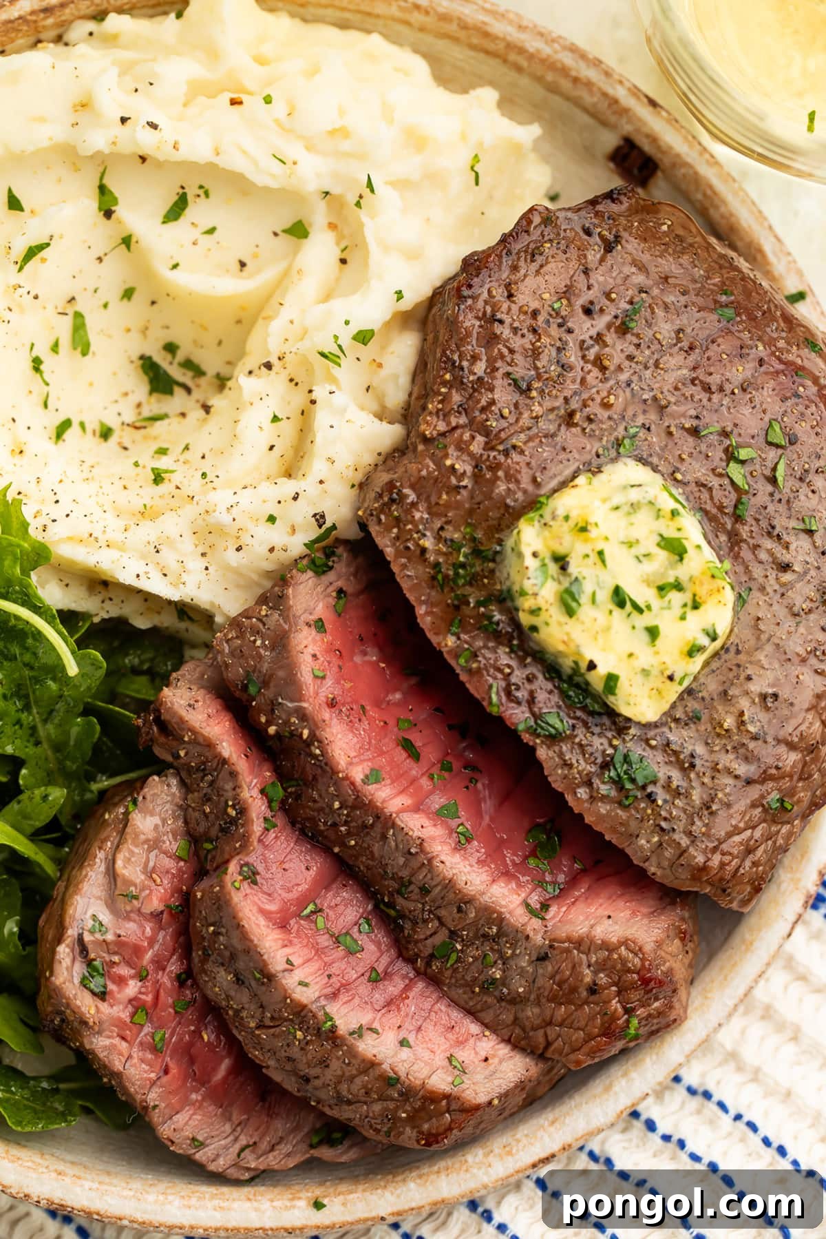 Close-up of medium-rare air fryer steak, topped with homemade herb butter, sliced and arranged on a plate with mashed potatoes and a small salad.