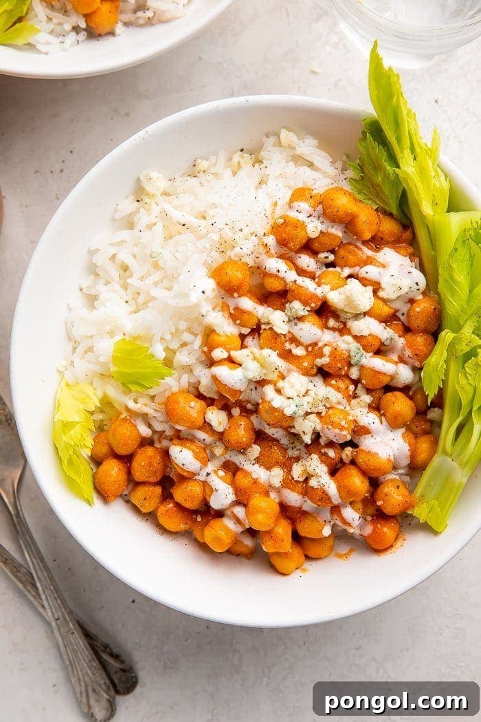 Fired Up Buffalo Chickpea Bowl 2 Creamy buffalo chickpeas generously coated in spicy sauce, served on a bed of white rice, garnished with fresh celery tops, blue cheese crumbles, and a drizzle of ranch dressing, creating a vibrant and satisfying meatless meal.