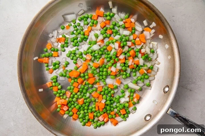 Mixed vegetables including diced onion, peas, and carrots, sizzling in a large silver skillet.