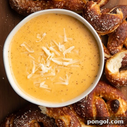 Overhead, top down look at a white bowl of rich yellow-orange beer cheese dip topped with shredded mozzarella surrounded by deep brown soft pretzels with salt.