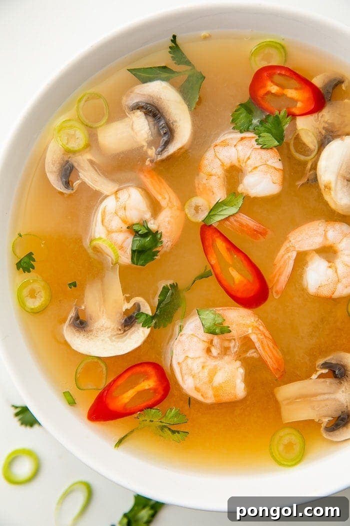 A steaming bowl of authentic Thai Tom Yum soup, generously filled with plump pink shrimp, vibrant red chili slices, tender mushrooms, and garnished with fresh green onion and bright cilantro leaves. The rich, clear broth promises a delightful balance of spicy, sour, and savory flavors, inviting you to a true taste of Thailand.