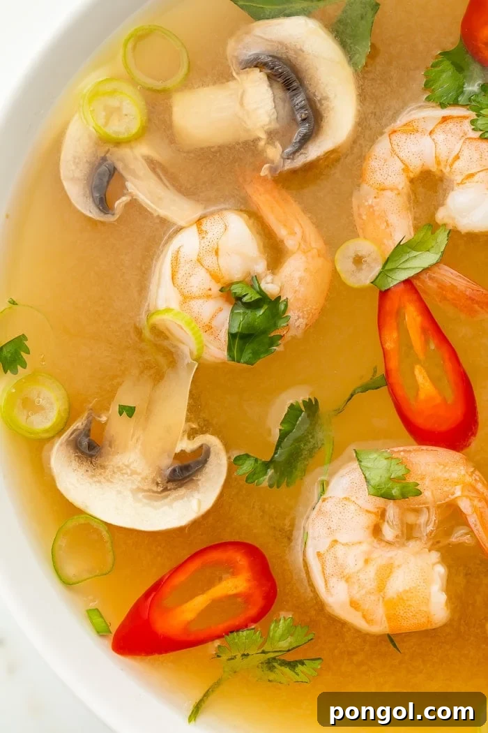 Close-up detailed view of an appetizing bowl of Tom Yum soup, showcasing the rich, clear broth. It features perfectly cooked, plump pink shrimp, vibrant red chili slices, tender sliced mushrooms, and a scattering of fresh green onions and bright green cilantro. The intricate textures and colors highlight the freshness and deliciousness of this classic Thai dish.