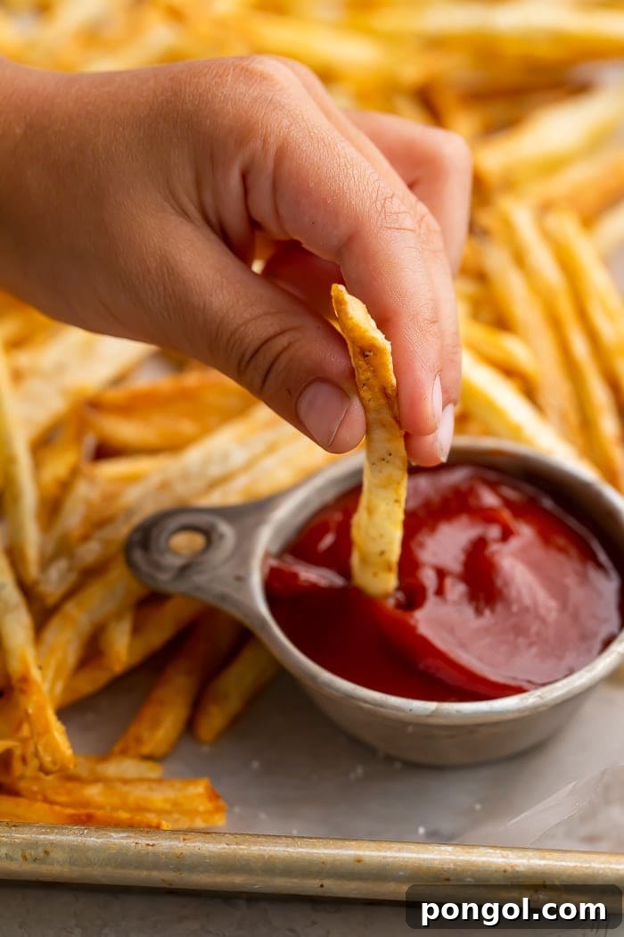 A child's hand reaches for a perfectly golden air fryer french fry, dipping it into a small white bowl of red ketchup.