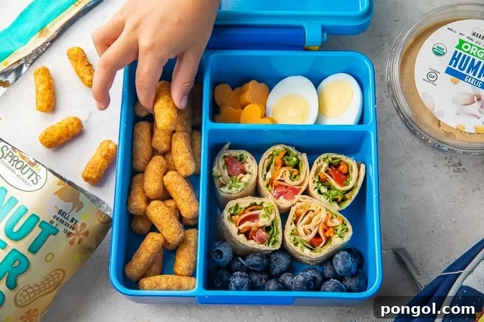 Child's hand reaching into a bento lunchbox filled with vegetarian snacks including hummus wraps, cheese, fruit, and peanut butter puffs.