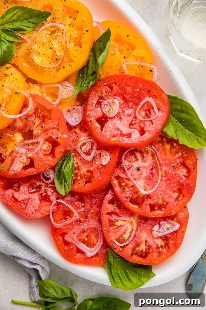 Rainbow Heirloom Tomato Medley 2 Vibrant yellow and red heirloom tomato slices artfully arranged on a white serving dish, generously adorned with thinly sliced shallots and fresh basil leaves.