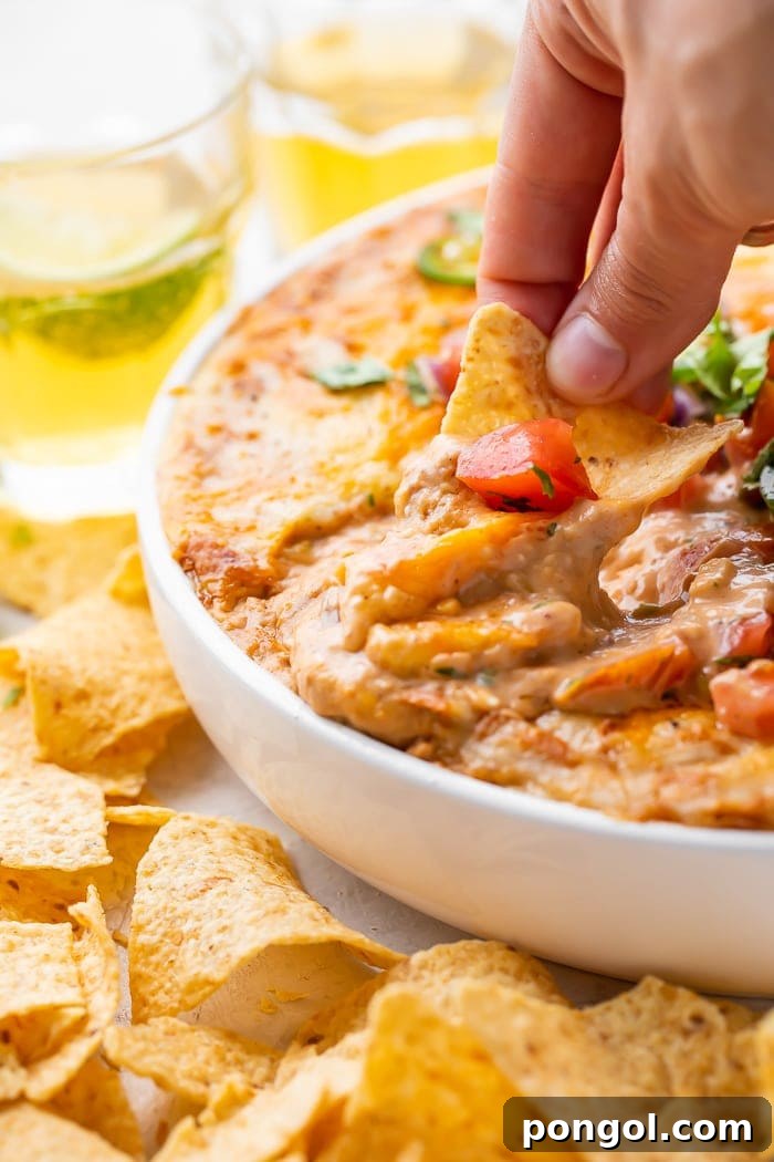 A hand dipping a corn chip into a bowl of bean dip in a bowl surrounded by glasses of beer and more chips