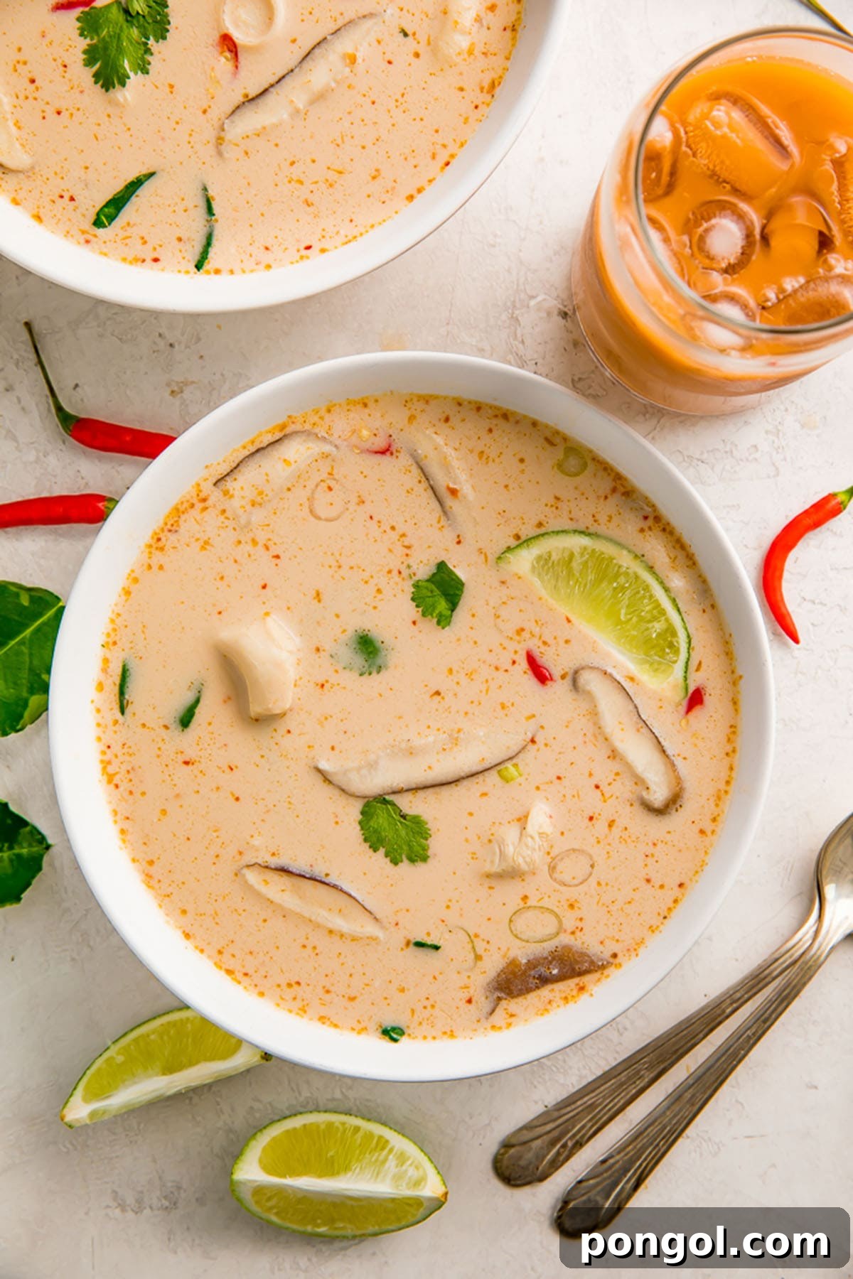 A bowl of creamy Crockpot Tom Kha Gai soup, garnished with fresh herbs, sits on a rustic table next to lime wedges and a glass of refreshing beverage.