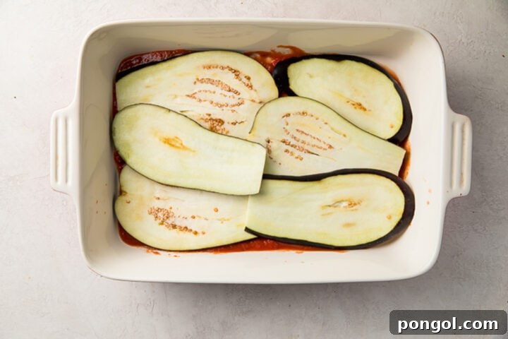 Sliced eggplants layered on top of a bed of bright red tangy marinara sauce in a white casserole dish.