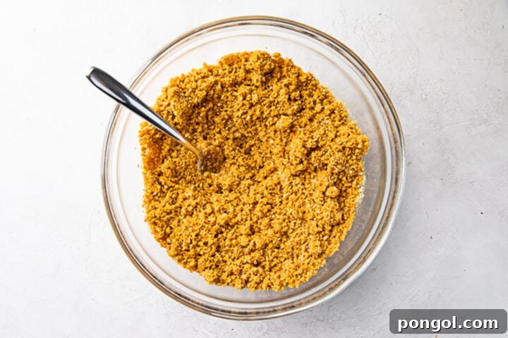 Graham cracker crust mixture in a large glass mixing bowl with a silver spoon, showing the perfectly moistened crumbs.