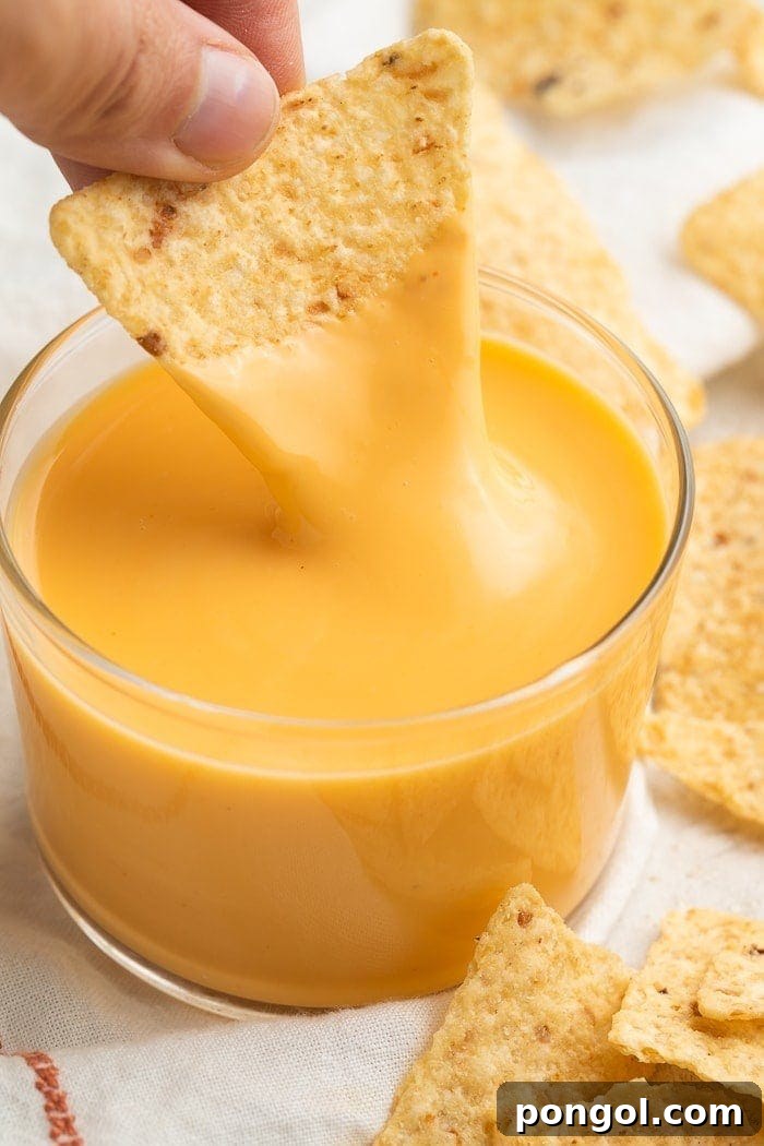 A triangular tortilla chip being dipped into a clear glass of Taco Bell nacho cheese sauce while surrounded by broken tortilla chips