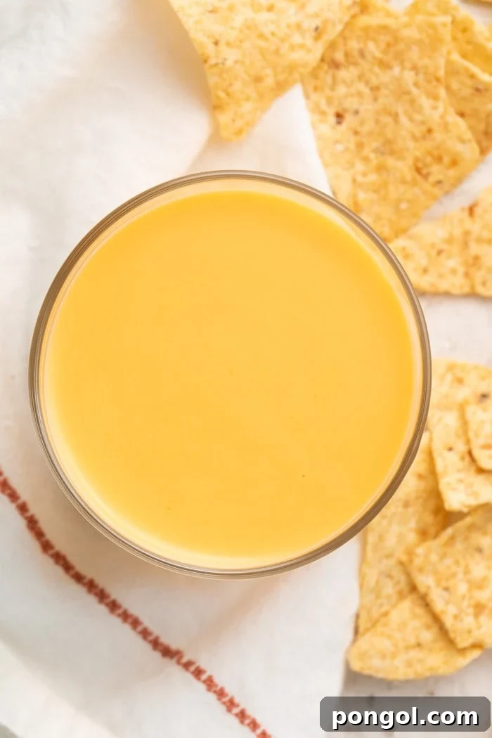 Overhead shot of a clear glass of Taco Bell nacho cheese sauce surrounded by a white napkin with a red line of embroidery and broken tortilla chips