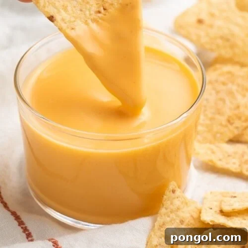 A triangular tortilla chip being dipped into a clear glass of Taco Bell nacho cheese sauce while surrounded by broken tortilla chips