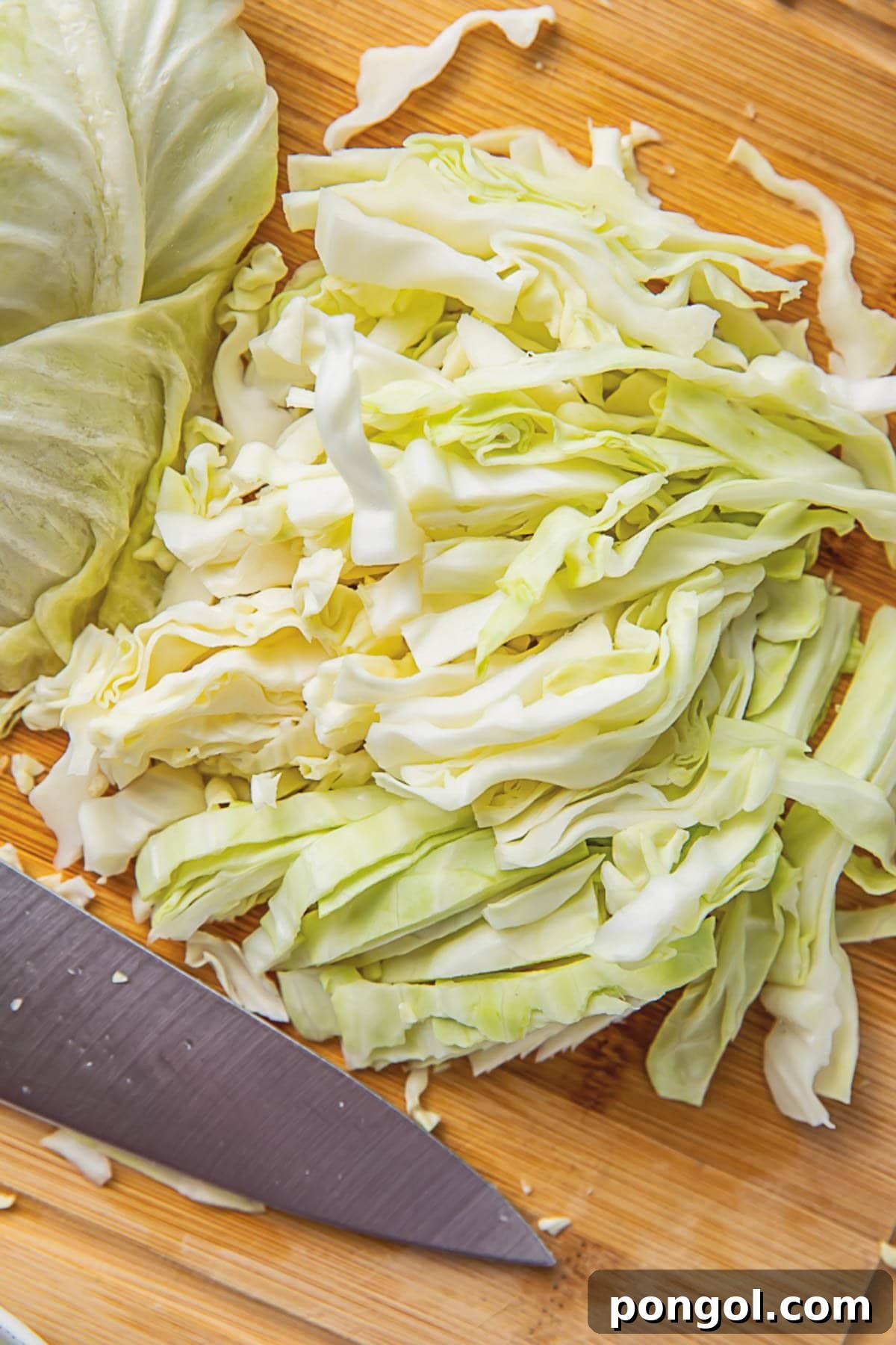 A head of cabbage, expertly shredded, resting on a rustic wooden cutting board next to a glistening sharp chef's knife.