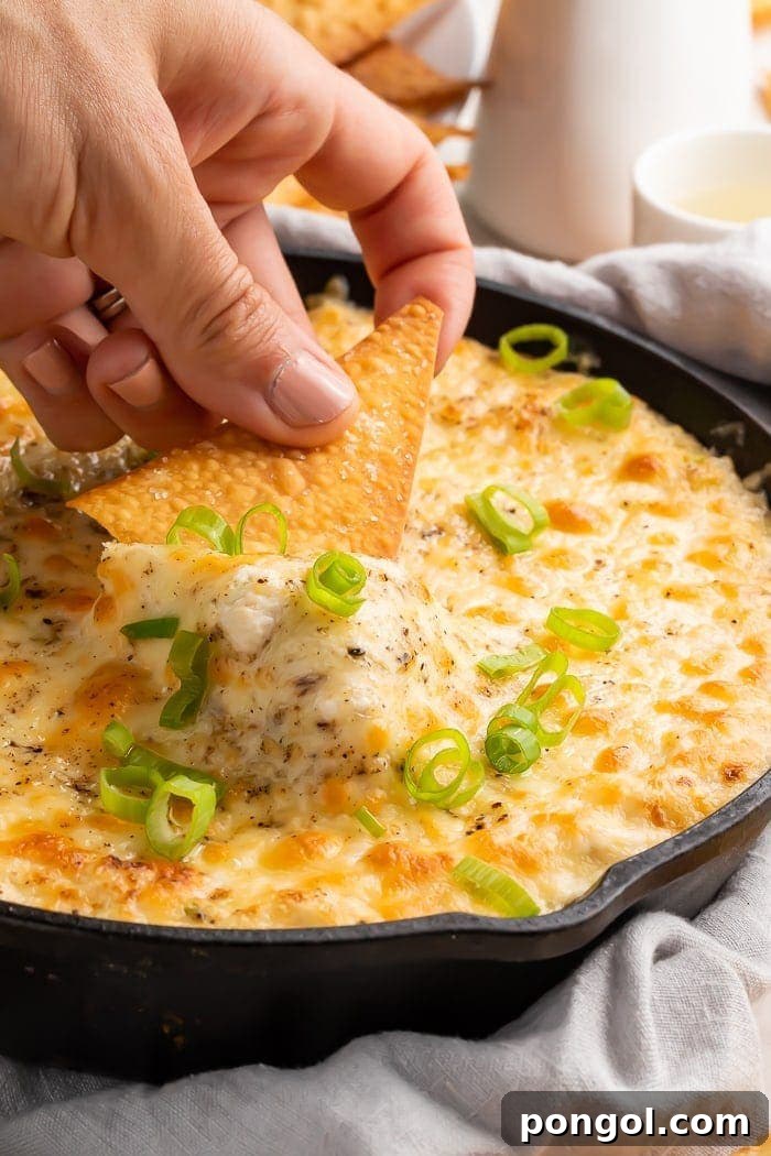 Deconstructed Crab Rangoon Dip 2 A hand dipping a chip into a cast iron skillet of crab rangoon dip, showcasing its creamy texture and golden-brown top