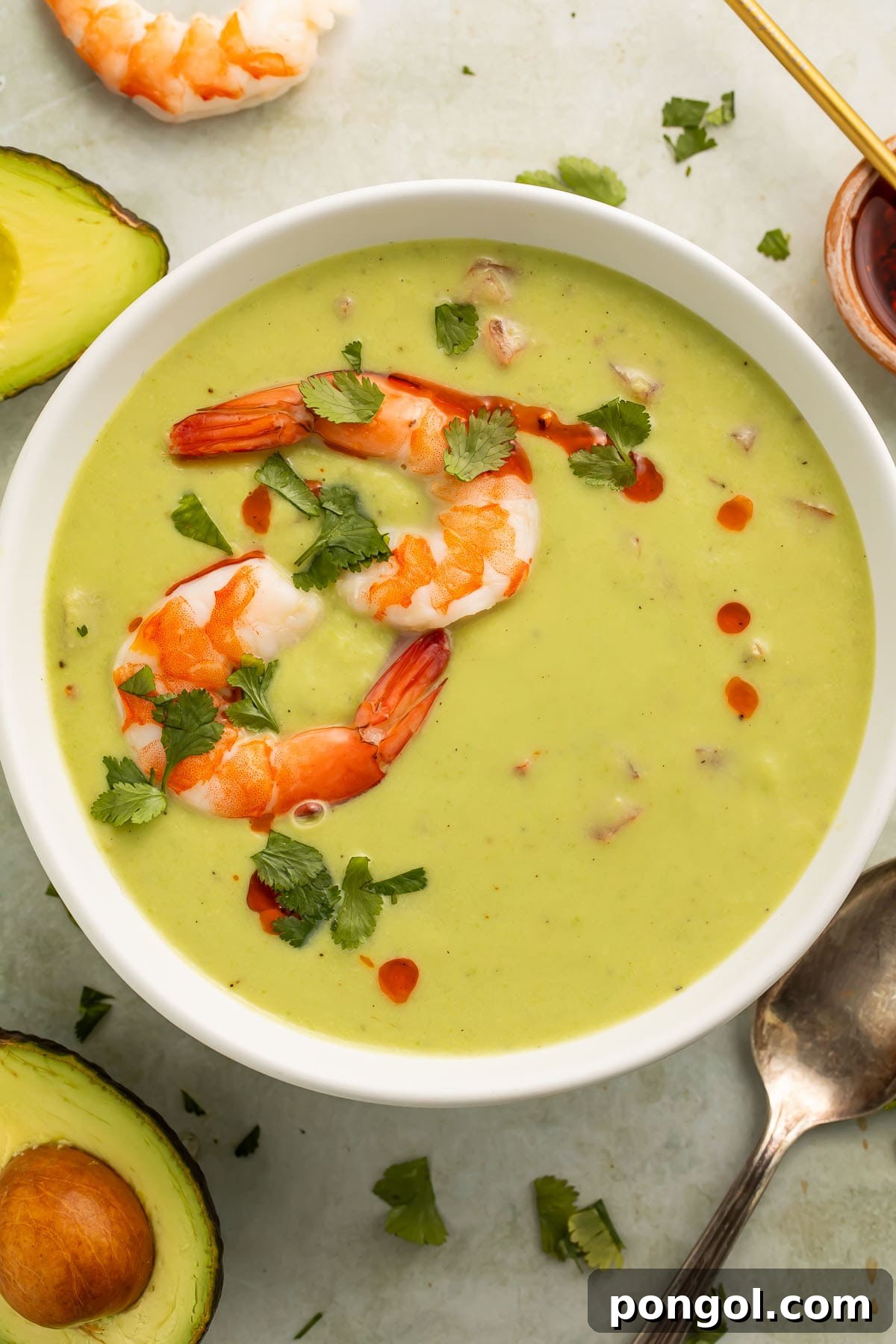 Velvety Chilled Avocado 2 A bowl of vibrant green chilled avocado soup, elegantly garnished with a swirl of crimson chili oil and two perfectly cooked pink shrimp, inviting a first delicious spoonful.