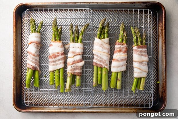 Uncooked asparagus spears wrapped in raw bacon laid perpendicularly on a wire rack over baking sheet