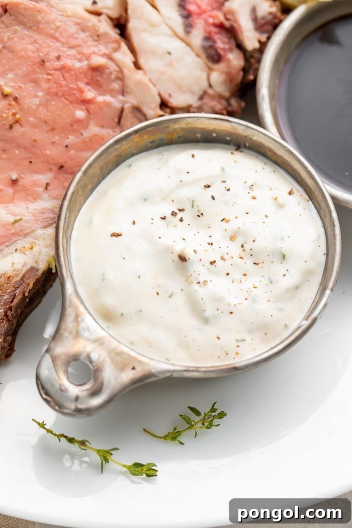 Close up of a silver dish with creamy horseradish sauce, showing its rich texture, next to slices of tender prime rib on a white plate.