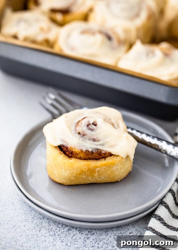 A single glazed Keto Cinnamon Roll on a blue plate, with a pan of more rolls in the background.