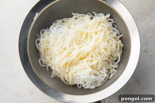 Boiled shirataki noodles in a silver mesh strainer over a silver bowl on a white counter