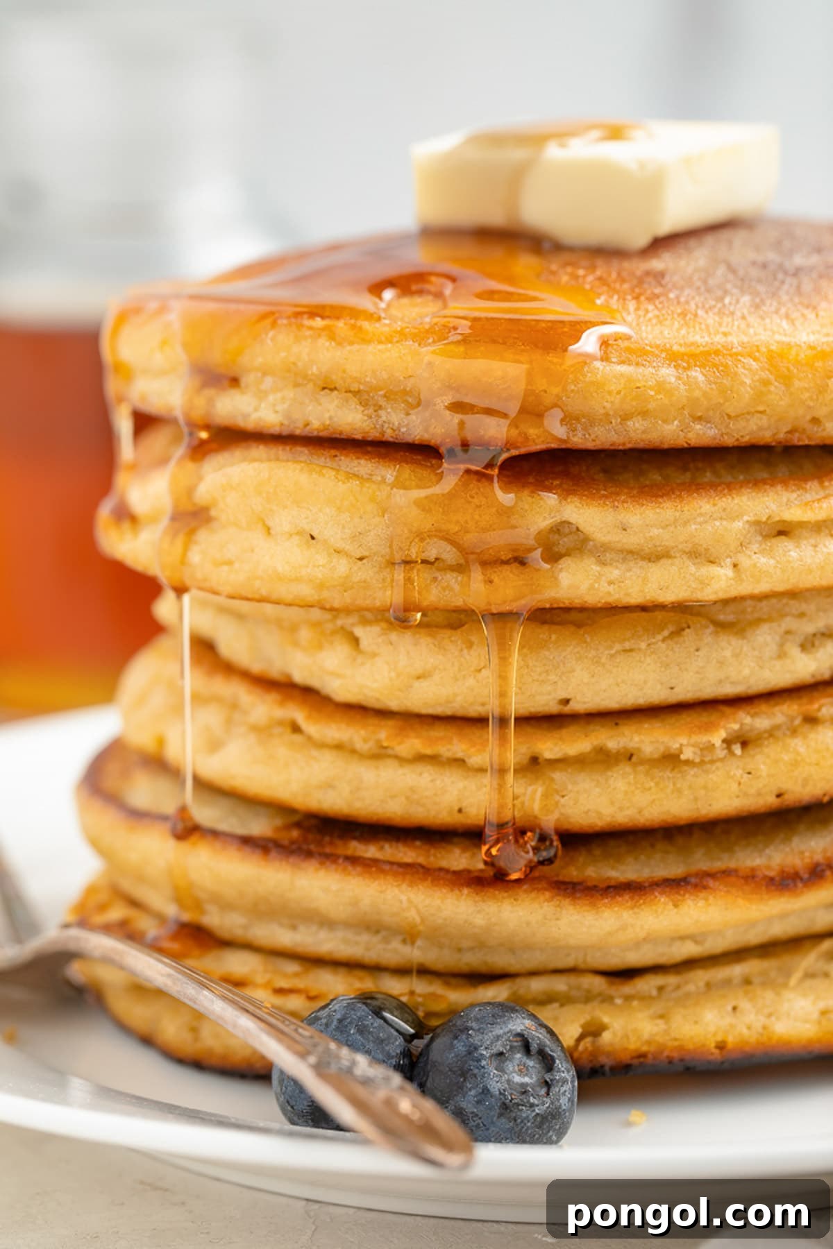 A stack of 6 paleo pancakes on a plate. 2 blueberries rest next to the pancakes, which drip with maple syrup.