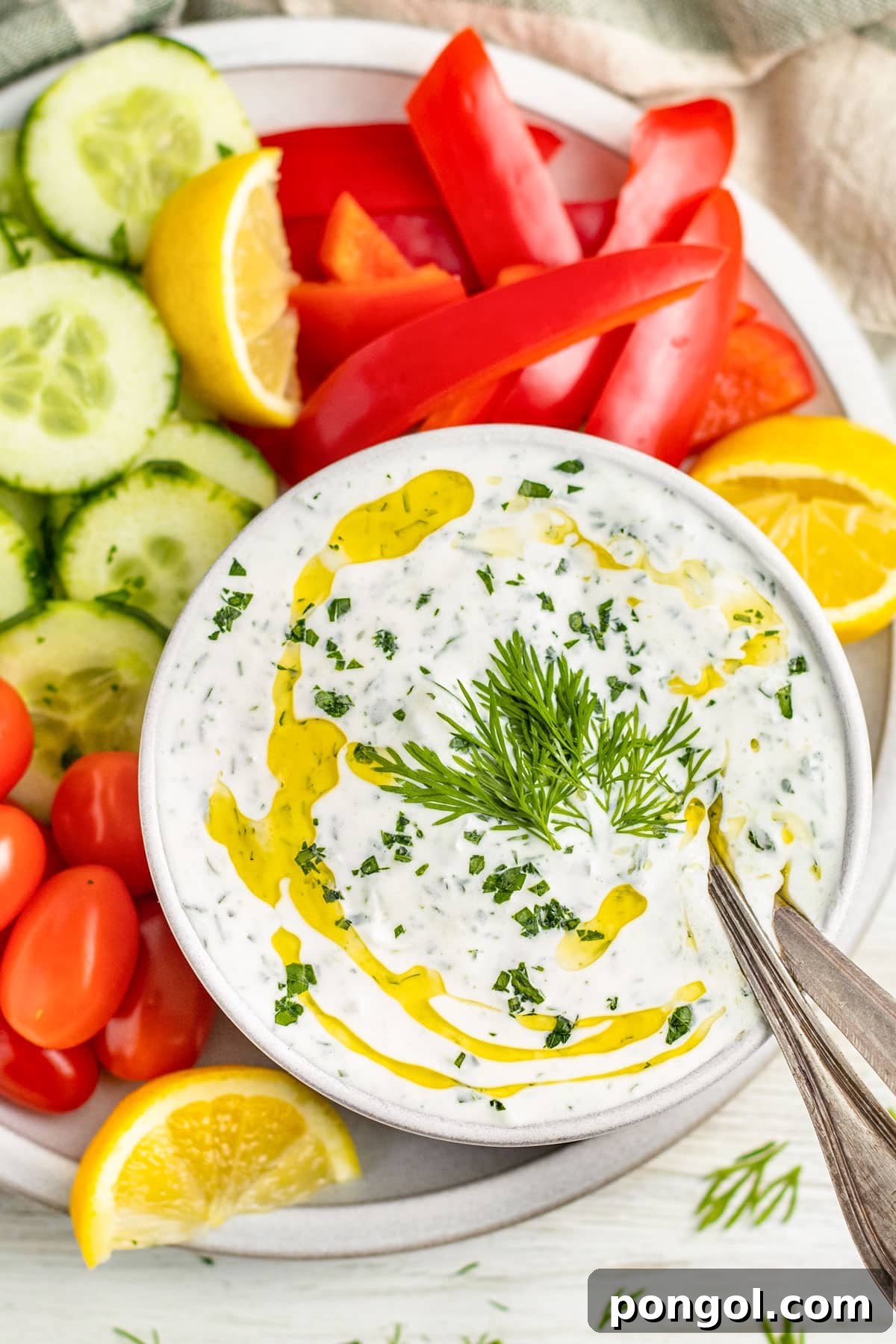 A bowl of creamy Whole30 tzatziki dotted with green fresh herbs and topped with a drizzle of olive oil, surrounded by cucumber slices, grape tomatoes, and bell pepper slices.