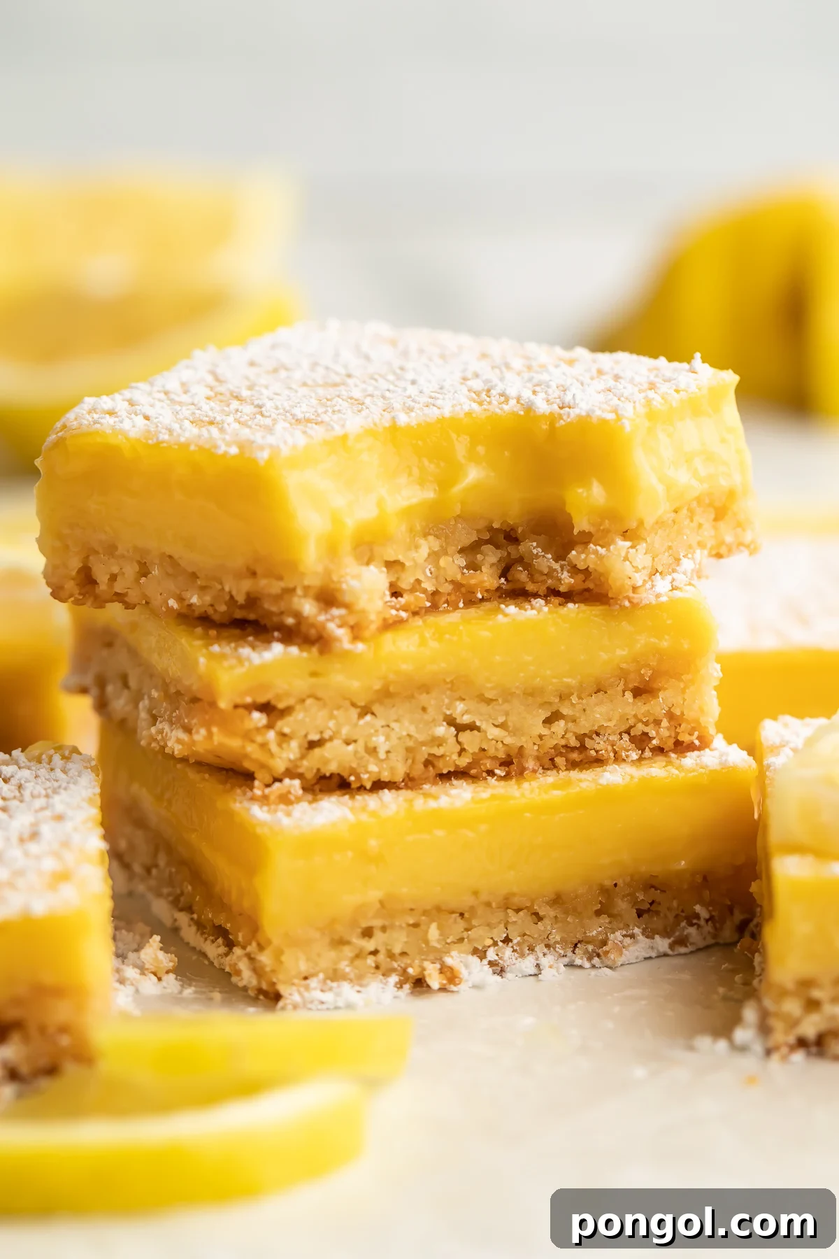 Three paleo lemon bars stacked on top of each other. A bite is missing from the corner of the lemon bar on top. Other lemon bars surround the stack, just out of focus.