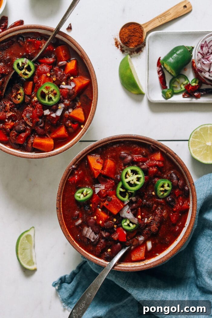 Two deep brown bowls holding a vibrant, deep red vegan chili, generously topped with sliced jalapeños and a silver spoon.