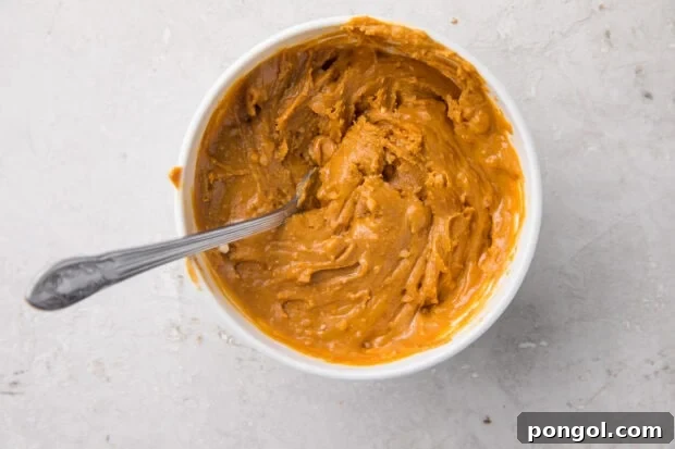 White mixing bowl holding peanut butter and powdered erythritol mixture with a silver spoon, demonstrating the smooth consistency prior to layering.