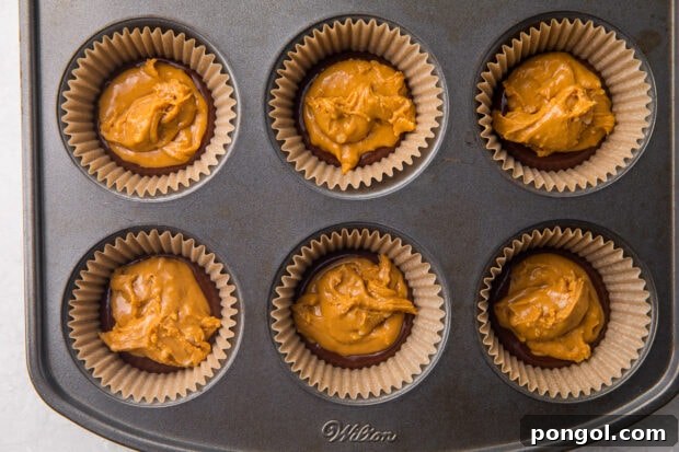 A precise dollop of peanut butter mixture being carefully placed on the hardened chocolate base within a cupcake liner.