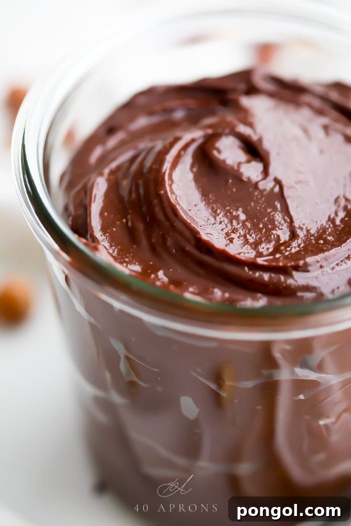 An intimate, close-up shot of creamy vegan Nutella in a small glass jar, highlighting its smooth, spreadable texture and rich chocolate-brown color. The focus is on the luxurious consistency, inviting viewers to imagine its taste.