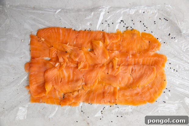 Delicately thinly sliced salmon meticulously arranged into a large, even rectangle on a sheet of plastic wrap adorned with sesame seeds
