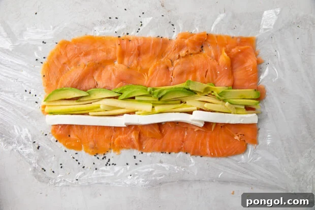 Precisely arranged cream cheese, creamy avocado, and crunchy cucumber layered atop the smoked salmon and sesame seeds on a sheet of plastic wrap