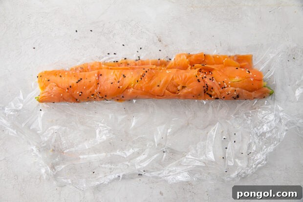 The smoked salmon sushi roll in progress, being expertly formed on plastic wrap, with the ingredients tightly encased