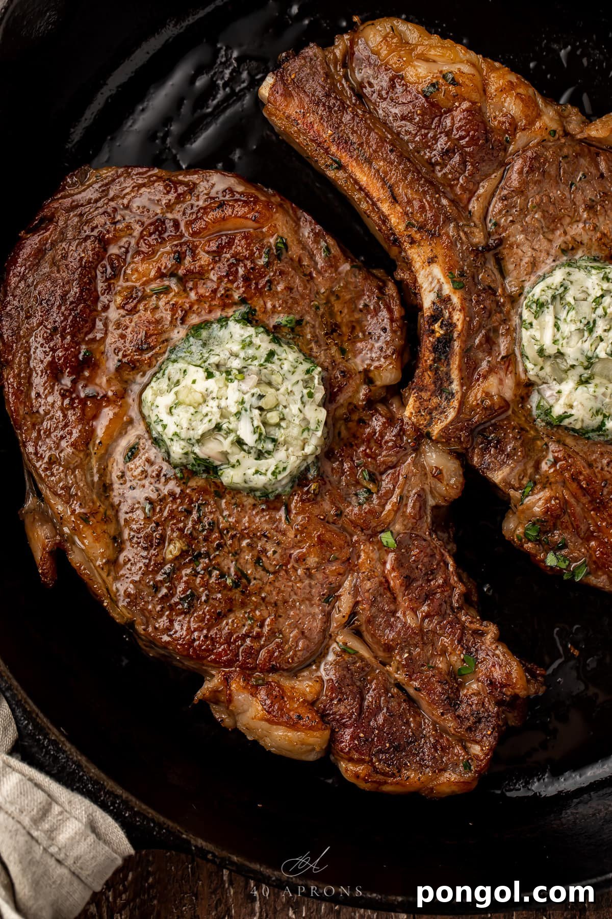 Two succulent bone-in ribeye steaks crowned with a generous dollop of melting garlic-herb butter, resting in a sizzling cast iron pan.