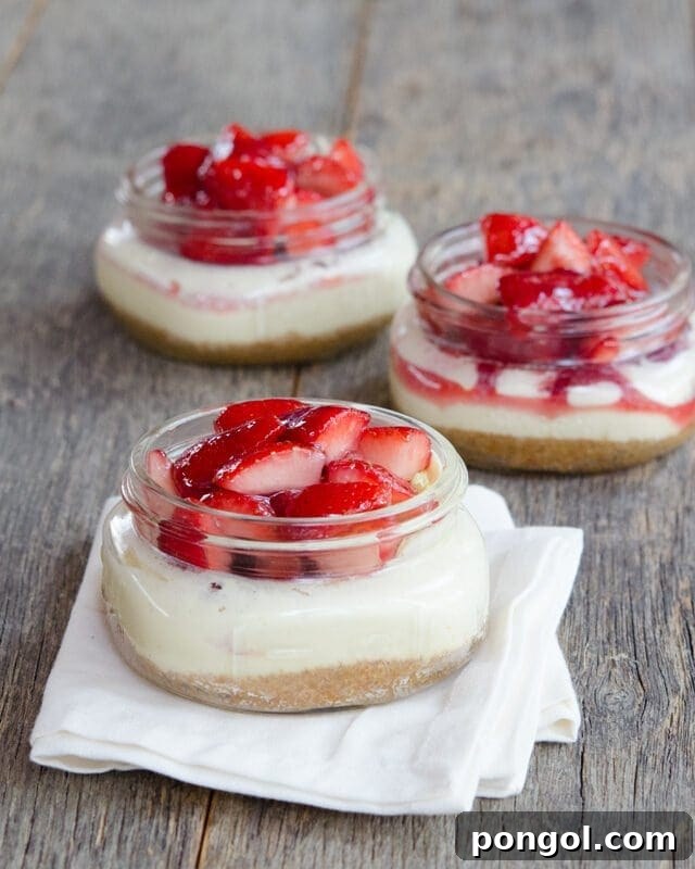Three charming mason jars filled with crust, creamy cheesecake, and fresh strawberries. The front jar rests delicately on a white napkin