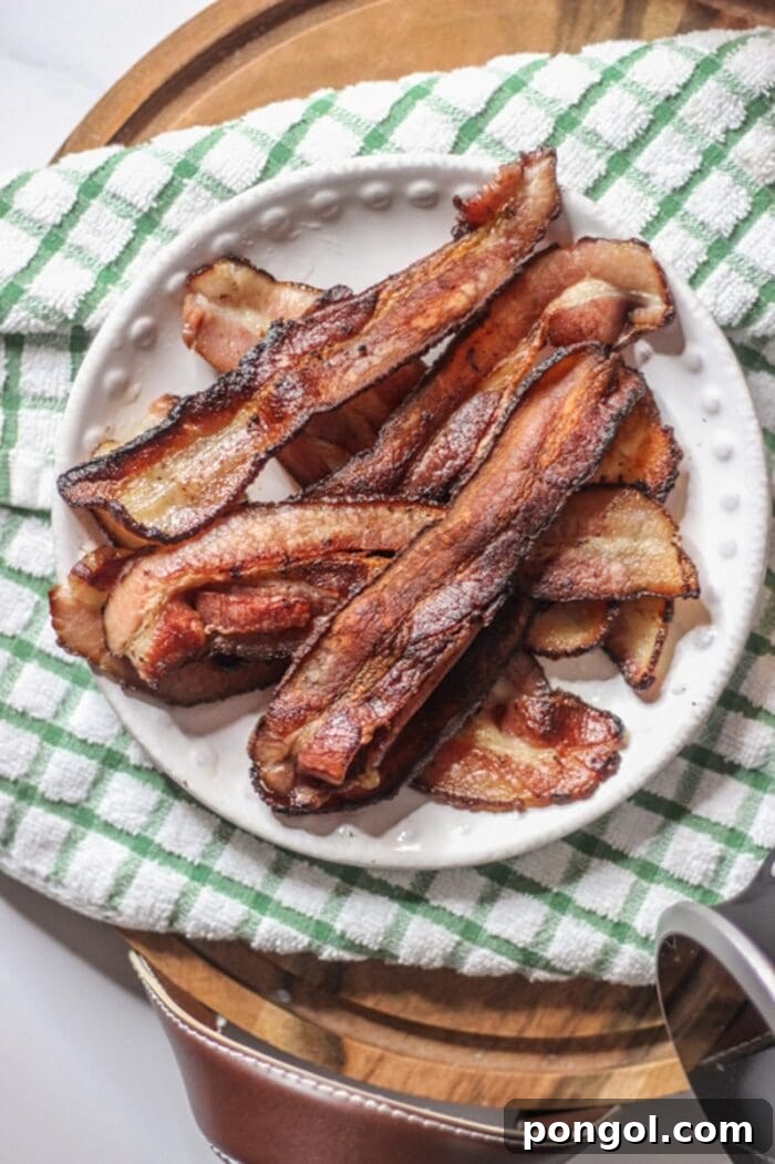 A generous plate of crispy, perfectly cooked bacon on top of a charming green and white checkered dish towel, resting on a wooden cutting board