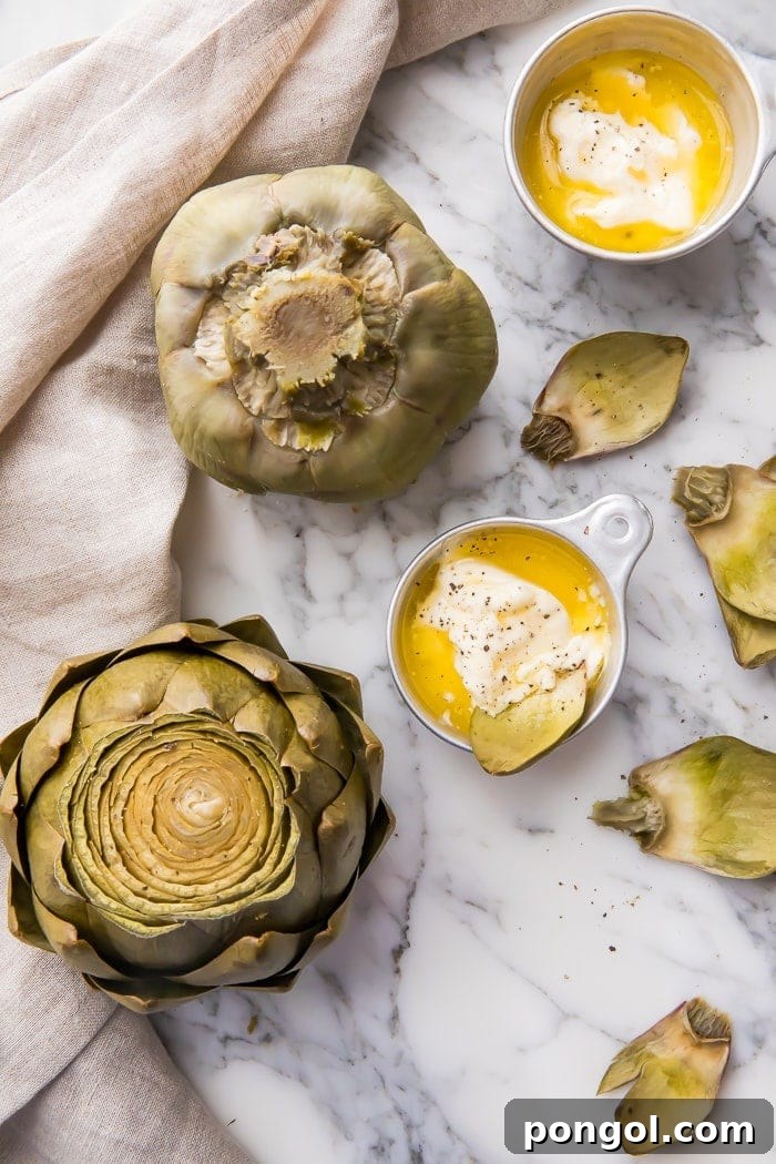 Two artichokes on a marble counter with bowls of dipping sauce