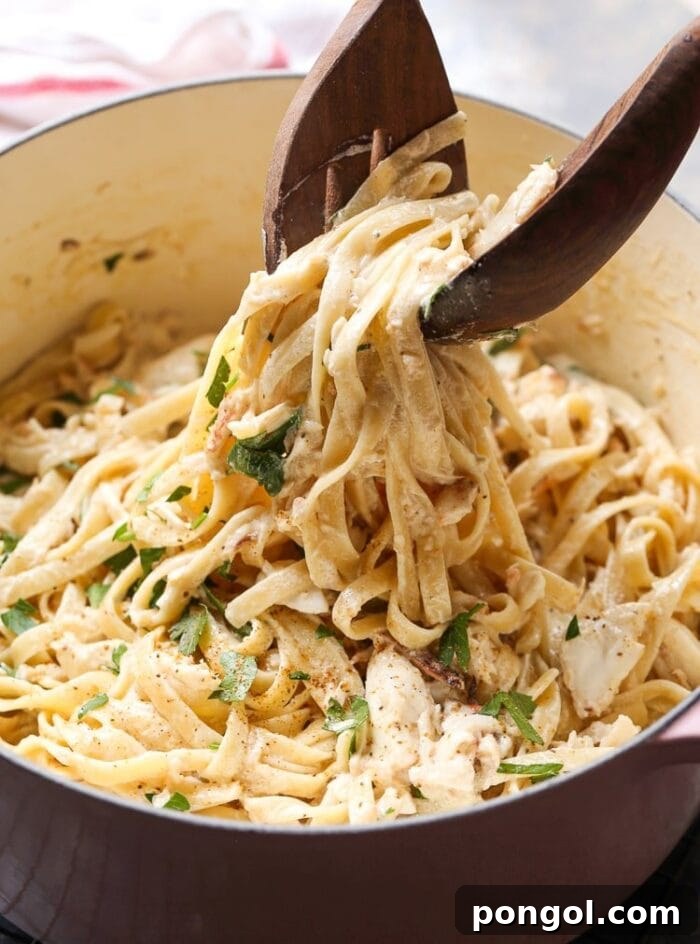 A large pot of creamy crab alfredo, with a scoop being lifted out by tongs, revealing rich pasta and crab.